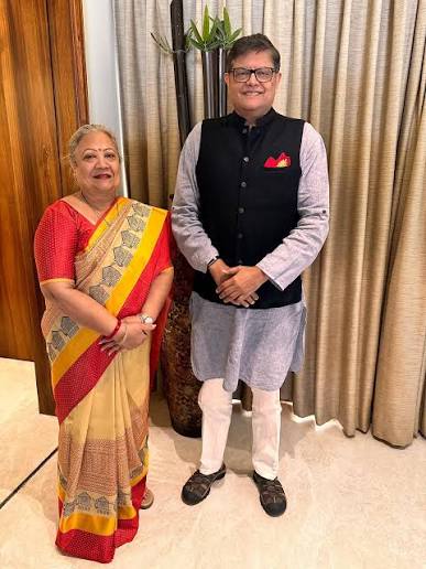 Baijayant Panda in a black waistcoat and grey kurta standing beside a woman in a red and yellow sari in a residence