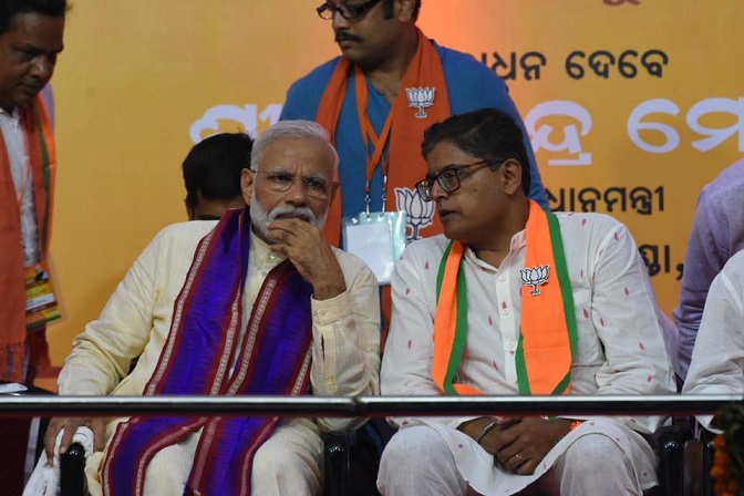 Baijayant Panda, in a white kurta and party stole, speaking to Prime Minister Narendra Modi as both sit on a stage in front of an Odia-language banner
