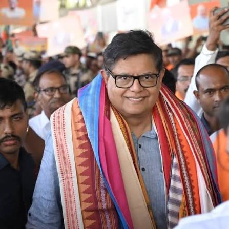 Baijayant Panda smiling, wearing black-rimmed glasses and a multi-coloured ceremonial stole, surrounded by a crowd at a public event