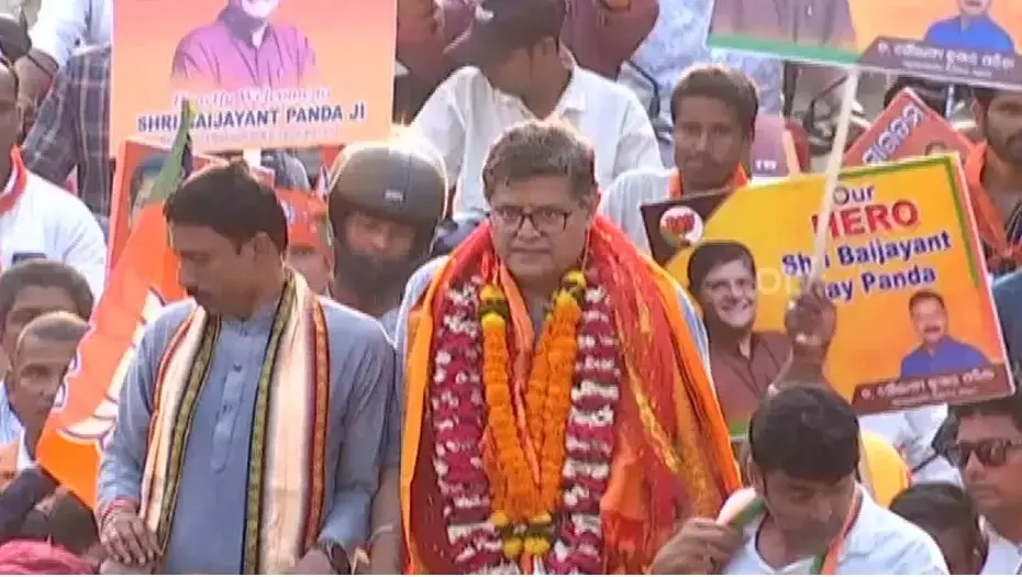 Baijayant Panda wearing marigold garlands and a stole at a crowded rally, with supporters holding placards bearing his name