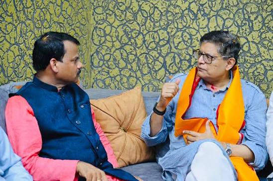 Baijayant Panda, wearing an orange stole and glasses, gestures while talking with a man in a blue waistcoat, both seated on a sofa